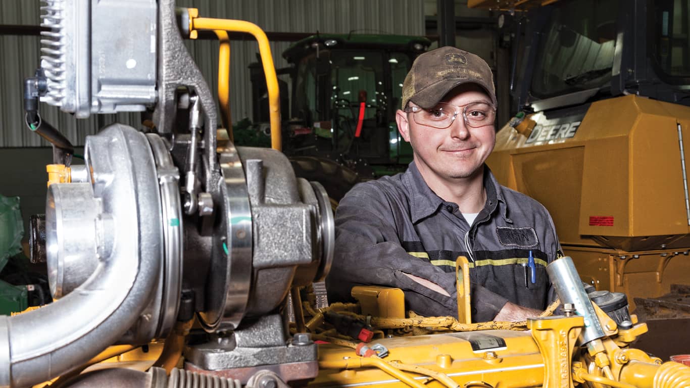 A man wearing safety goggles smiling next to a John Deere engine